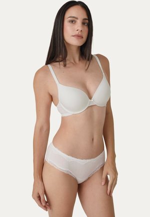 Frau trägt weißen gepolsterten BH und Unterwäsche mit Spitzensaum, steht vor hellem, einfarbigem Hintergrund und blickt leicht nach links.