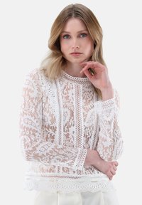 Blusa bianca in pizzo con maniche lunghe, caratterizzata da motivi floreali intricati, orlo a scallop e bordature dettagliate lungo il collo e i polsini.