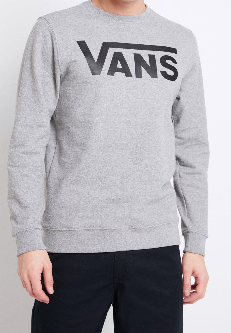 Personne portant un sweat-shirt gris clair à manches longues avec un grand logo noir "VANS" sur la poitrine et un pantalon noir.