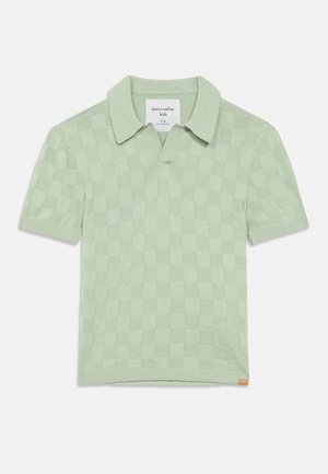 Πλεκτό παιδικό πόλο με κοντά μανίκια σε ανοιχτό πράσινο χρώμα, με καρό σχέδιο και μικρή δερμάτινη ετικέτα μάρκας στο κάτω τελείωμα.