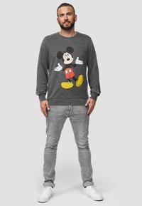 Šedá mikina s velkým tištěným obrázkem Mickey Mouse v červené a žluté barvě, doplněná o světle šedé jeansy a bílé tenisky.