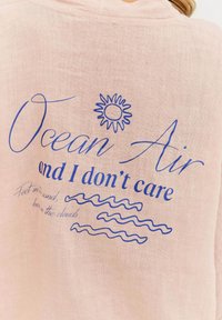 Svetloružová tkanina s modrým textom. Text znie: "Oceánový vzduch a je mi to jedno." Obsahuje grafiku slnka a vzory vĺn.