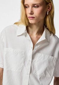 Lyshåret kvinde med sølvringøreringe og en hvid kortærmet skjorte med to brystlommer, der ser let nedad og mod venstre.