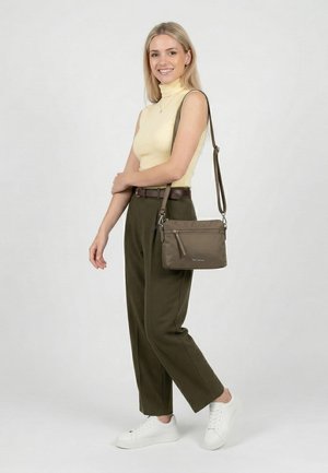 Lächelnde Frau in gelbem ärmellosem Oberteil und olivgrüner Hose steht, trägt eine dunkelbraune Umhängetasche und weiße Sneaker vor weißem Hintergrund.