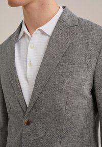 Ljusgrå blazer med fiskbensmönster, notched lapels och en enda brun knapp. Bärs över en vit texturerad polo skjorta.