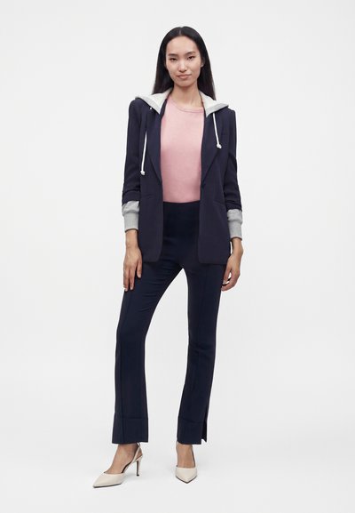 Jeune femme portant un blazer bleu marine avec des manches grises de sweat à capuche, un haut rose, un pantalon bleu marine et des talons hauts beiges, debout devant un fond blanc uni.