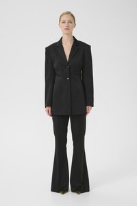 Čierny prispôsobený blejzer s výraznými ramenami, dvoma gombíkmi a štruktúrovanými švami, kombinovaný s rozšírenými čiernymi nohavicami a špicatými žltými lodičkami.