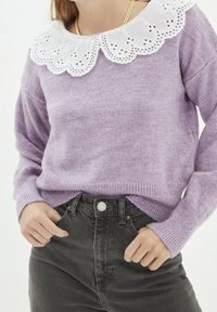 Maglione viola con maniche lunghe e colletto in pizzo bianco a scallop. Indossato con jeans neri a vita alta, con dettagli di bottoni e tasche.