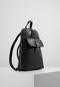 Mochila de cuero negra con bolsillo frontal y cierre de cremallera, de pie sobre una superficie blanca con un fondo claro y liso.
