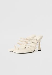 Des sandales blanches à talons aiguilles présentent plusieurs lanières sur le dessus, une forme de pied carré et un accent métallique sur le talon. Texture lisse.