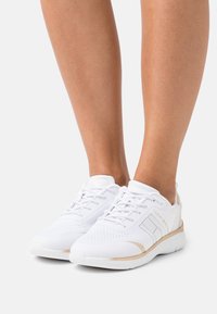 Chaussures de sport blanches avec une tige en mesh texturé, dotées d'une fermeture à lacets et d'accents dorés sur la semelle intermédiaire. Semelle extérieure en caoutchouc lisse.