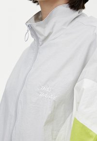 Giacca grigia chiaro con colletto alto, caratterizzata da una cerniera frontale, accenti bianchi e un dettaglio verde neon sul maniche. Il testo riporta "TRÈS TRENDY."