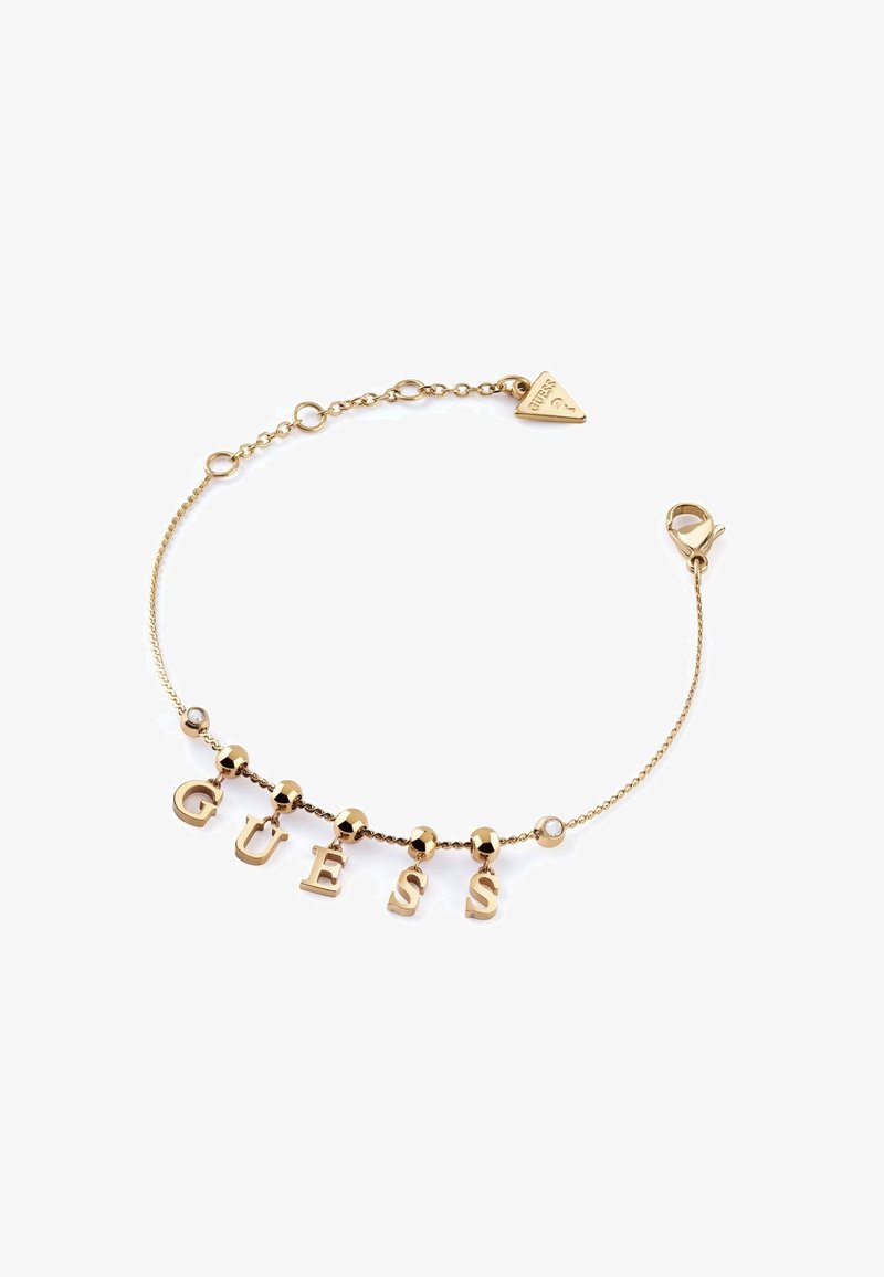 Bracelet en chaîne dorée avec le mot "GUESS" composé de lettres suspendues, des perles rondes et une étiquette en forme de triangle. Fermeture par fermoir.