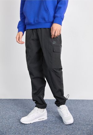 Person trägt schwarze Nike Cargohosen, weiße Sneaker und einen blauen Sweatshirt und steht auf einem grauen Teppich vor einem einfarbigen Hintergrund.