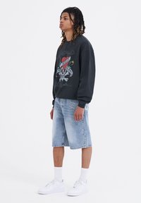 Svart sweatshirt med grafisk design, ljusblå denimshorts och vita sneakers. Modellen står på sidan och visar upp en avslappnad passform och casual stil.