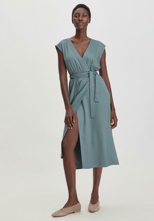 Kleid aus weichem, hellgrünem Stoff mit tiefem V-Ausschnitt, kurzen Ärmeln, einem Bindegürtel in der Taille und einem seitlichen Schlitz bis zum Oberschenkel. Flache, beigefarbene Schuhe.