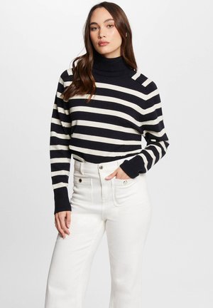 Pull en maille à col roulé avec des rayures horizontales bleu marine et crème, à manches longues et poignets côtelés, associé à un jeans blanc taille haute.