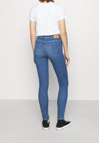 Vaqueros skinny de tiro alto en azul, hechos de denim, con dos bolsillos traseros y una parche de cuero marrón. Combinados con zapatillas negras.