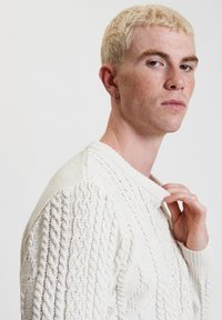 Pullover in maglia color crema con pattern a treccia sui maniche e colletto a coste, indossato da una mano che regola il scollo.
