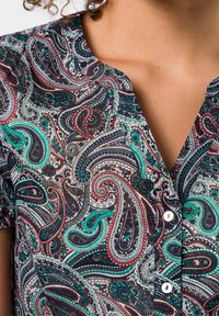 Blouse à manches courtes présentant un motif paisley en rouge, vert et bleu sur un fond sombre, avec un devant à bouton et un col en V.