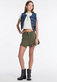 Denim vest, witte cropped top, olijfgroene rok met gerimpelde zoom en grijze enkellaarzen met gespsluiting. Eenvoudige, casual outfit.