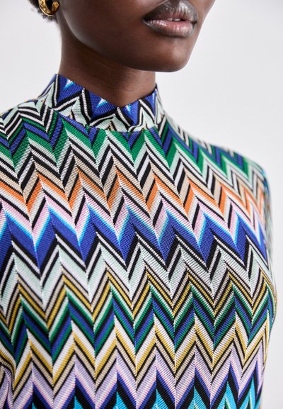 Top colorato con collo alto, caratterizzato da un motivo a zigzag nei colori verde, blu, arancione e nero. Il tessuto sembra testurizzato con strisce vivaci e contrastanti.