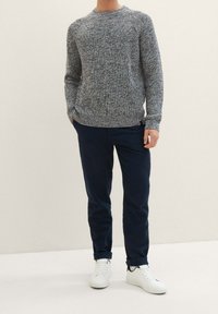 Man in een grijze gebreide trui, donkerblauwe opgerolde broek en witte sneakers die met één hand in zijn zak staat tegen een eentonige achtergrond.