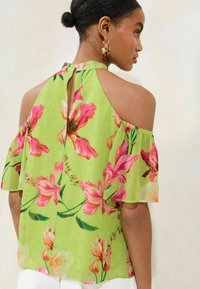Blusa a maniche corte di un brillante verde con motivi floreali in rosa e arancione. Presenta una schiena aperta con un dettaglio a chiave. Tessuto liscio e leggero.