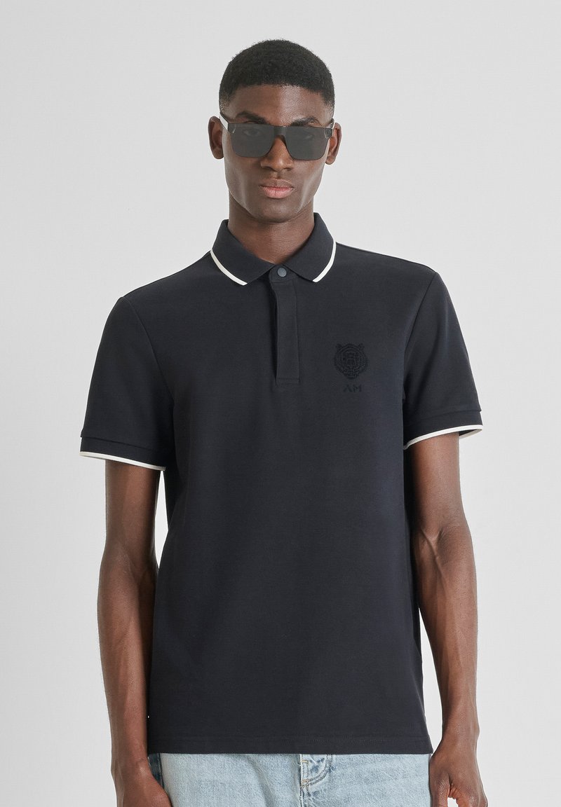 Crna polo majica s bijelim ovratnikom i naglascima na rukavima. Ima vezeni logo na prsima i dugmad na zapvaku. Glatka tekstura.