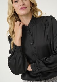 Blusa di satin nero con colletto arricciato, chiusura con bottoni e maniche a sbuffo. Caratterizzata da una texture liscia e una leggera lucentezza.