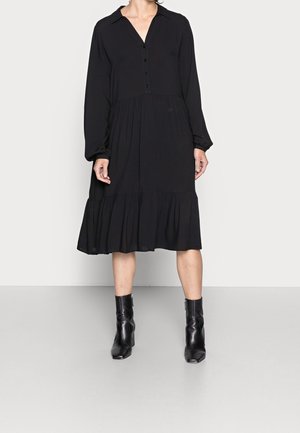 Femme portant une robe midi noire à manches longues avec des boutons et un ourlet à volants, associée à des bottines noires, debout sur un fond uni.