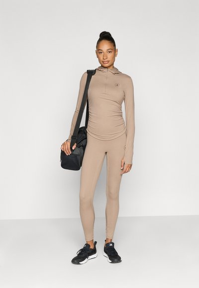 Beige athletic outfit consisting of a fitted long-sleeve top and leggings, made of smooth fabric, with a hood and subtle logo details. Black gym bag.