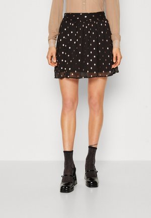 Woman wearing black skirt with metallic dots, beige blouse, glittery black socks, and black patent leather heels standing on plain white floor.