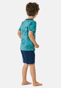 Turquoise t-shirt met voetbalpatronen, korte mouwen en een marinekleurige kraag, gecombineerd met donkerblauwe shorts, op blote voeten, gezien van achteren.