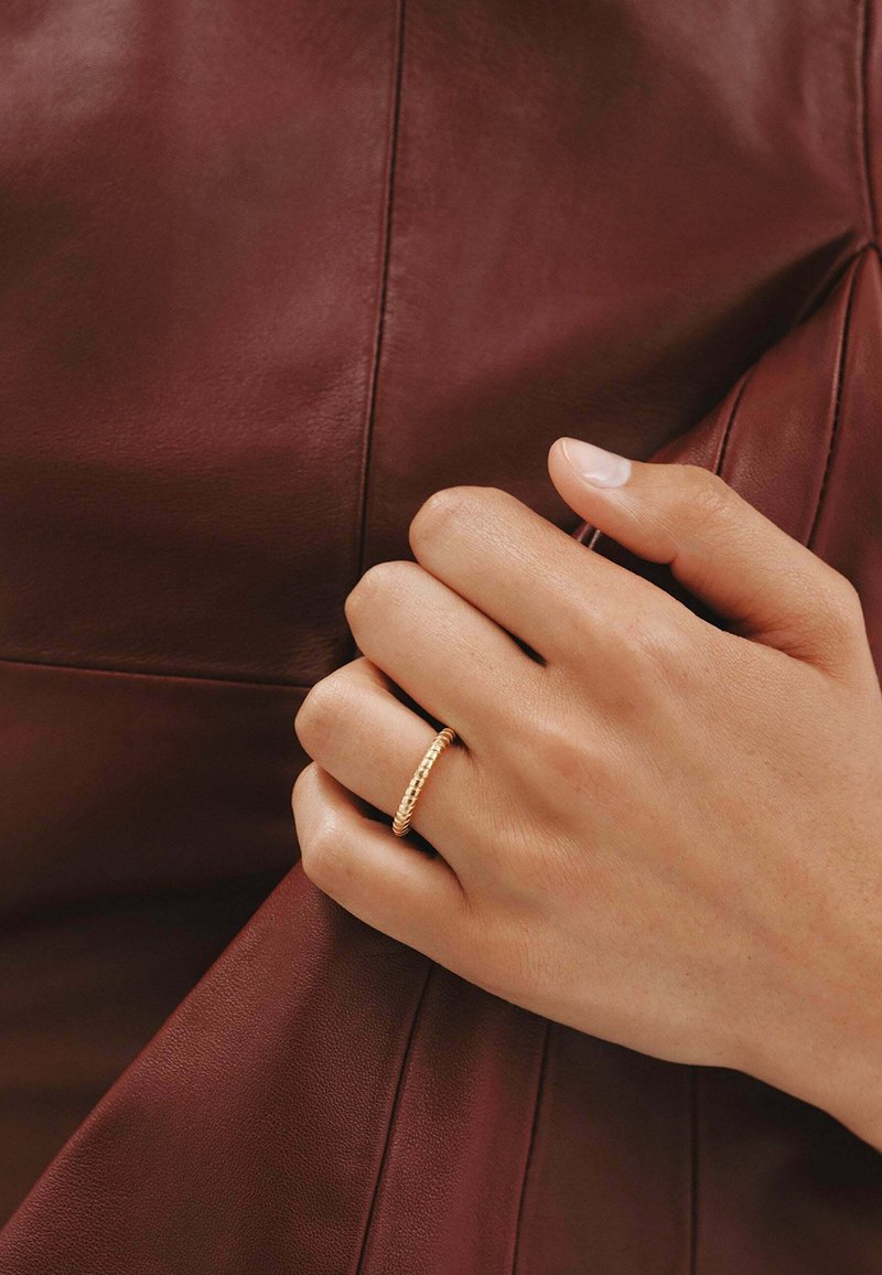 Anillo de oro con un diseño retorcido y texturizado, llevado en una mano que sostiene una prenda de cuero burdeos, mostrando piel suave y uñas limpias.