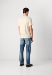 Young man standing facing away wearing a cream short-sleeve shirt, blue faded jeans, and black sneakers on a plain white background.