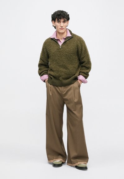 Olivgrüner strukturierter Pullover mit Reißverschlusskragen über einem hellrosa Hemd mit Kragen. Lockere braune Hosen und grüne und schwarze Schuhe runden das Outfit ab.