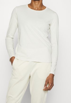 Persona vestida con una camiseta blanca ajustada de manga larga con cuello redondo y pantalones de color claro, de pie con una mano en el bolsillo.