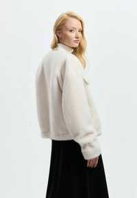 Blondynka w białym puszystym żakiecie i czarnej spódnicy spogląda przez ramię na tle jednolitego białego tła.