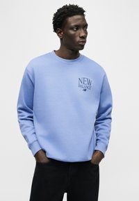 Νέος άνδρας που φοράει ένα ανοιχτό μπλε φούτερ New Balance με τα χέρια στις τσέπες, κοιτάζοντας πλάγια μπροστά σε ένα απλό φωτεινό φόντο.