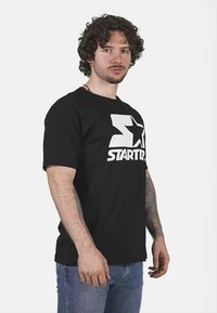 T-shirt in cotone nero con un grande logo bianco "START" e grafica a stella. Maniche corte e vestibilità rilassata. Indossato con jeans blu.