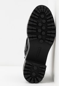 Chaussure en cuir noir avec une semelle en caoutchouc robuste présentant des crampons hexagonaux. Détail de fermeture éclair latérale visible, ainsi que l'étiquette de taille 37.