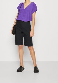 Lila blus med kort ärm och V-ringning samt samlad detalj, i kombination med svarta knälånga shorts och svarta sko som är lätta att ta på. Svart axelremsväska.