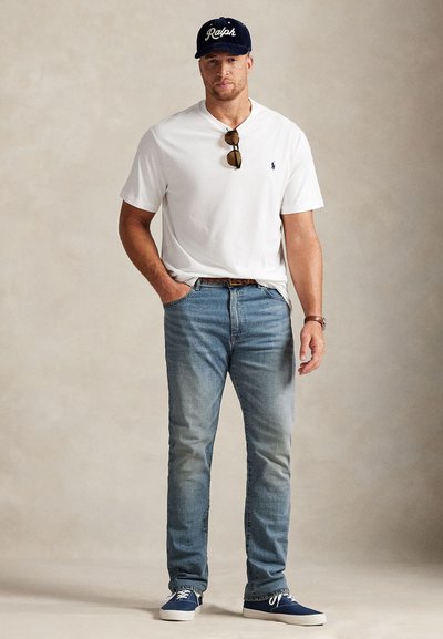 Bílé bavlněné tričko, světle modré džíny, tmavě modré tenisky a tmavě modrá kšiltovka s bílým nápisem; sluneční brýle zavěšené na tričku.