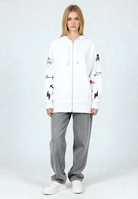 Felpa bianca con cappuccio e cerniera, con stampe grafiche colorate sulle maniche. Abbinata a jeans grigi a gamba larga e sneakers bianche.