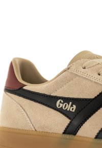 Deportivas de gamuza beige con acentos negros, talón burdeos y suela de goma clara. Presenta detalles de logo y elementos de diseño cosidos.