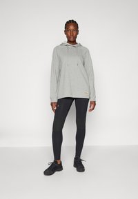 Grijze hoodie met trekkoorden en logo, gecombineerd met zwarte leggings met textuur op de knieën. Model draagt zwarte sportieve schoenen.
