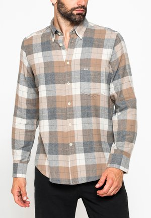 Uomo con camicia a quadri beige, grigia e bianca con taschino sul petto, in piedi con le mani vicino alla vita, su sfondo bianco.