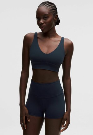 Donna con i capelli intrecciati che indossa un reggiseno sportivo scuro e pantaloncini aderenti a vita alta, in piedi contro uno sfondo semplice.