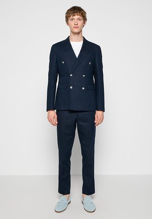 Giovane uomo in piedi che indossa un blazer doppiopetto blu navy, pantaloni abbinati, camicia bianca e mocassini azzurri su uno sfondo grigio semplice.